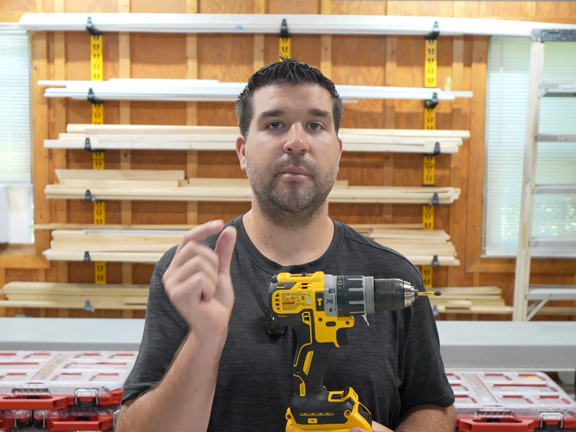 Jeremy in his workshop holding up a cordless drill, with the lumber rack and tool boxes behind him.